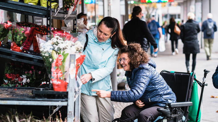 Zwei Frauen, davon eine im Rollstuhl, schauen sich die Angebote eines Blumenladens an.