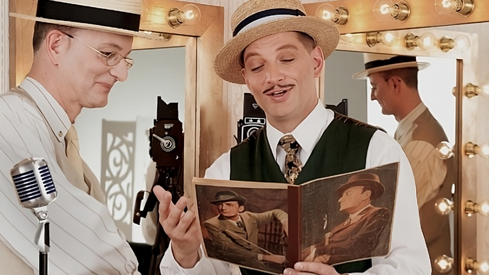 Foto: Die Wilden Witwer, zwei mittelalte weiße Männer mit Pork Pie-Hützen und zwanziger-Jahre-garderobe lächelnd in einem Buch blätternd