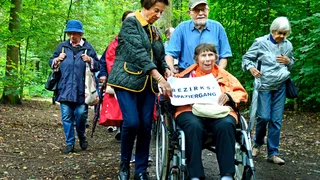 Foto: Eine Spaziergangsgruppe im Wald, im Bildvordergrund eine Dame im Rollstuhl