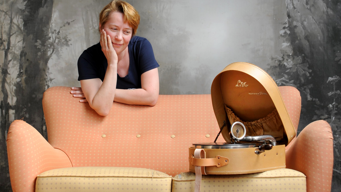 Foto: Karen Freis, eine weiße Frau mittleren Altern, stützt sich auf die Rückenlehne eines nostalgisch wirkenden apricotfarbenden Sofas und schaut verträumt auf ein Reisgrammophon, das auf der Sitzfläche steht