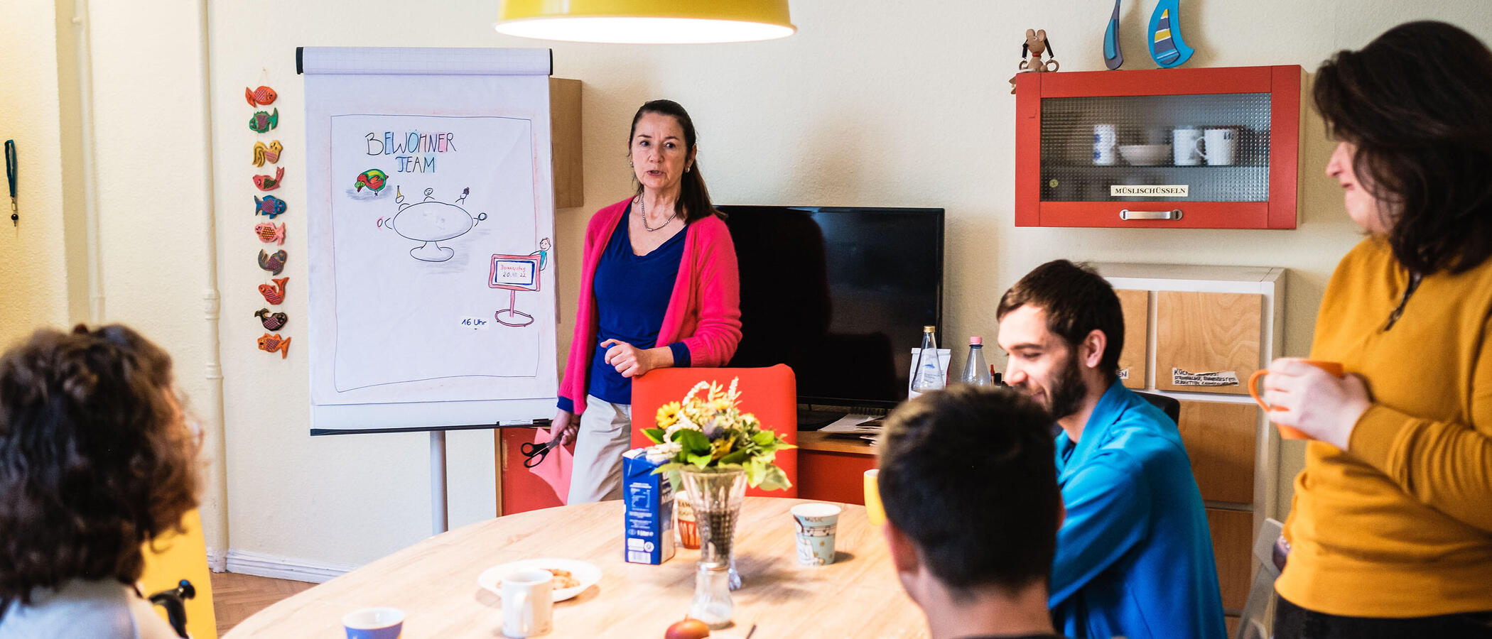 Mehrere Person sitzen an einem runden Tisch. Eine Frau steht neben einem Flipchart und erklärt etwas.