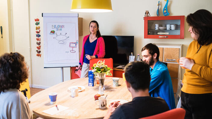 Mehrere Person sitzen an einem runden Tisch. Eine Frau steht neben einem Flipchart und erklärt etwas.