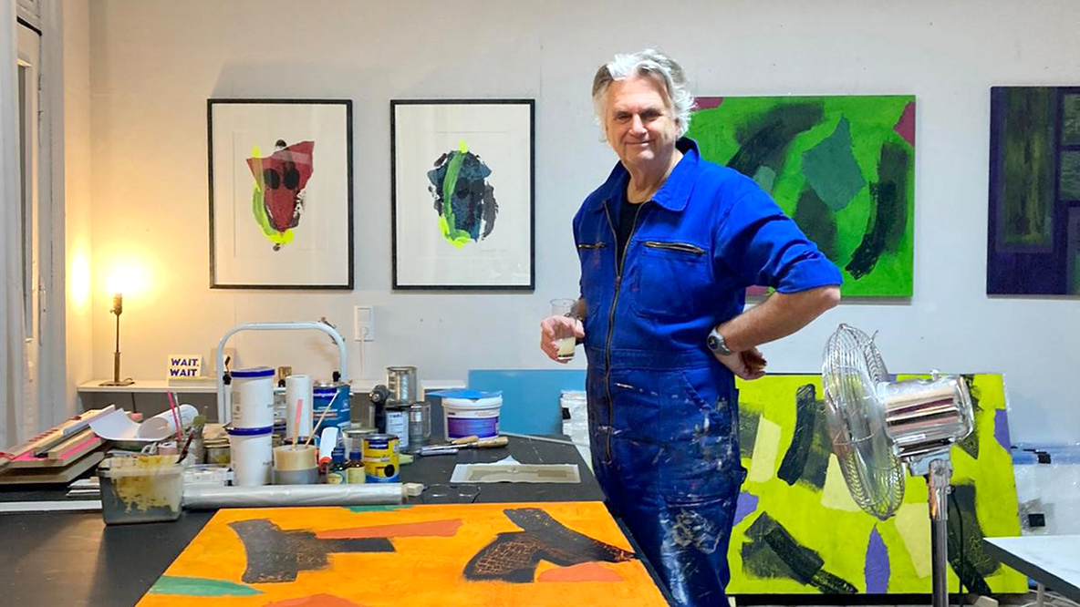 Foto: Klaus Decker, ein Mann im blauen Overall mit weißen Harren mittleren Alters, lächelnd in seinem Atelier, im Hintergrund Bilder an den Wänden