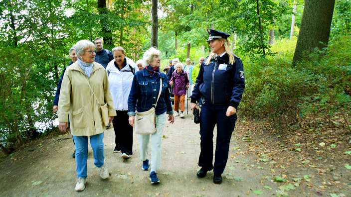 Foto: Eine gemischte Gruppe älterer Damen und Herren beim Spaziergang unter Bäumen, recht im Bild eine Polizistin in Uniform