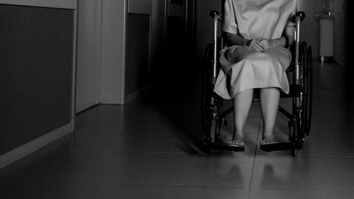 Foto: Eine Frau im Rollstuhl in Schwarzweiß, zu sehen bis zur Schulter, sie steht auf einem leeren Krankenhausflur