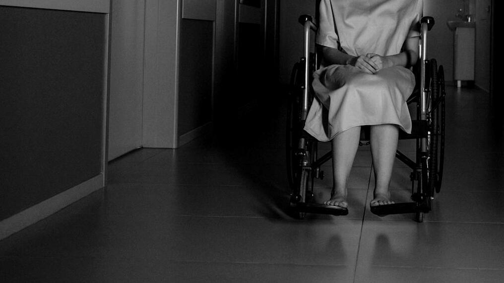Foto: Eine Frau im Rollstuhl in Schwarzweiß, zu sehen bis zur Schulter, sie steht auf einem leeren Krankenhausflur