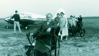 Foto: Historisches Bild aus ca. 1960er Jahren, es zeigt einen reiferen Rollstuhlfahrer in einem älteren Rollstuhl, im Hoimntergrund ein Mann vor einem Sportflugzeug und zwei der Zeit entsprechend modisch gekleideten Damen