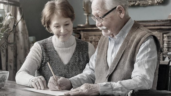 Foto: Ein Paar im Seniorenalter sitzt in einem altertümlich anmutendem Wohnzimmer an einem Eichentisch; der Mann im Rollstuhl schreibt etwas auf einen Zettel, die Frau zu seiner Rechten lächelt