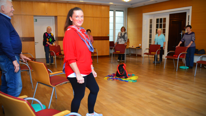 Foto: Sechs Frauen und ein Mann machen Übungen der Sturzprävention, hinter einem Stuhl stehend; im Vordergrund die lachende Trainerin in sportlicher Kleidung, in der Raummitte diverse Trainingsutensilien 