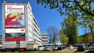 Fotocollage: Das Umbauquartier der Villa Donnersmarck vor blauem Himmel, im Vordergrund geparkte Autos, an der Häserwand des funtionellen mehrgeschossigen Gebäudes die Vorderseite des neuen Programmheftes 1/2026