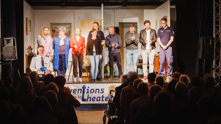 Foto: Blick aus dem Publikum auf die Bühne des Präventionstheaters der Polizei Abschnitt2; auf der Bühne das diverse Ensemble der Polizei, zwei Personen in Uniform