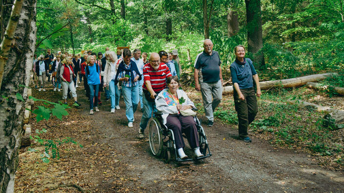 Foto: Eine bunt gemischte Spaziergangsgruppe im Wald, im Vordergrund ein älteres Ehepaar mit Rollstuhl, er schiebt sie