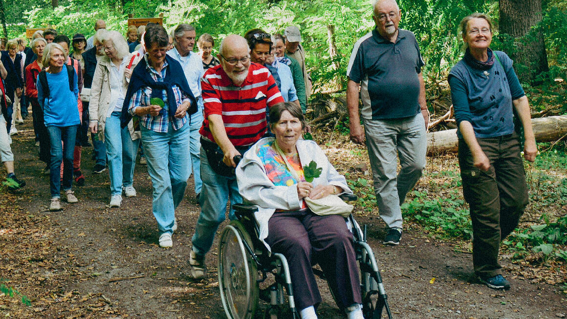 Foto: Eine bunt gemischte Spaziergangsgruppe im Wald, im Vordergrund ein älteres Ehepaar mit Rollstuhl, er schiebt sie