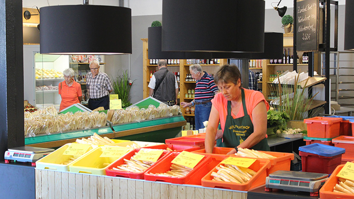 Foto: Eine Dame sotiert Spargel in einem Hofladen, im Hintergrund weitere Kundschaft