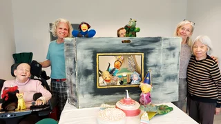 Foto: Die Kunstgruppe der Villa Donnersmarck, ein Mann und vier Frauen, lächelnd vor der Kulisse für ihr Puppentheater; die Dame links im Bild nutzt einen Rollstuhl