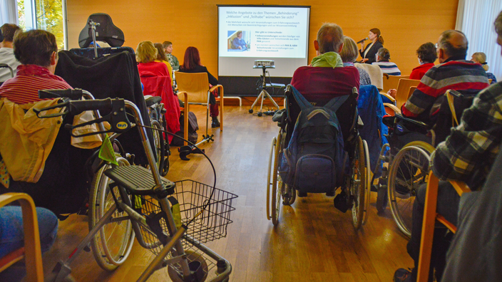 Foto: Blick in den Saal bei einer Diskussionsveranstaltung, im Vordergrund im Publikum mehrere Personen im Rollstuhl mit dem Rücken zum Betrachtenden