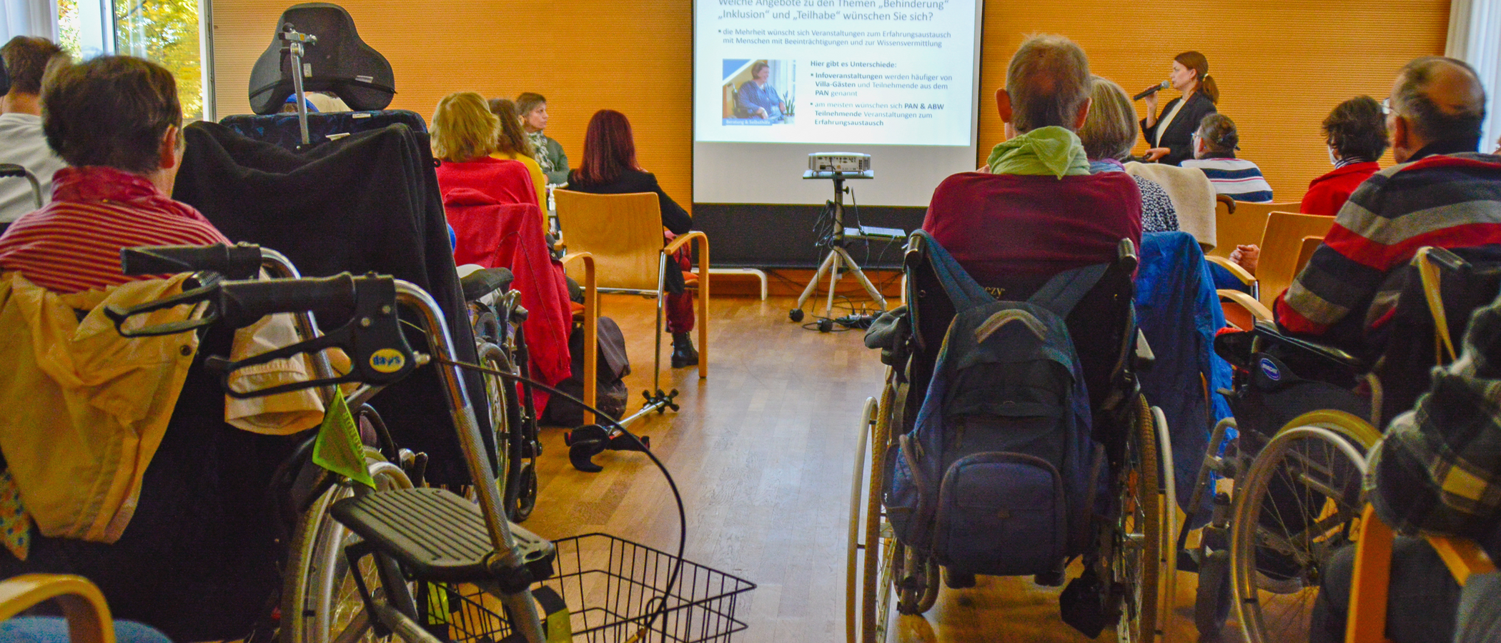 Foto: Blick in den Saal bei einer Diskussionsveranstaltung, im Vordergrund im Publikum mehrere Personen im Rollstuhl mit dem Rücken zum Betrachtenden