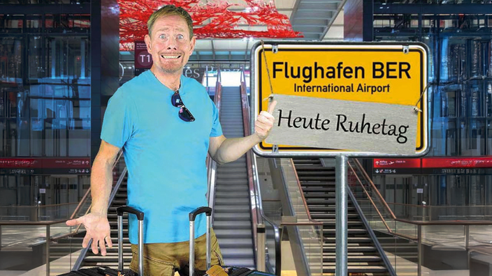 Foto: Donato Plögert, ein weißer Mann mittleren alters, steht vor zwei Rollkoffern vor einer Rolltreppe am Flughafen; mit einer Grimasse zeigt er auf ein gelbes Ortschild, auf dem "Flughafen Berlin, heute Ruhetag" steht