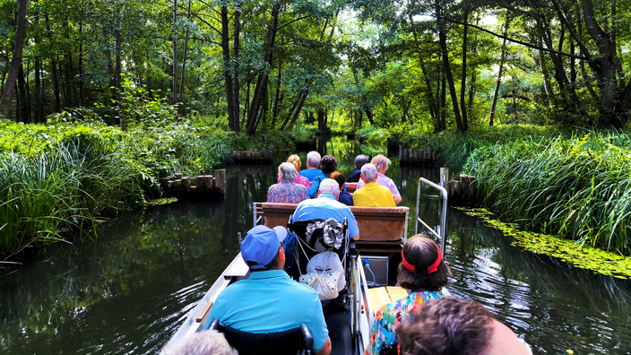 Foto: Eine diverse Reisegruppe, fotografiert von hinten, auf einem Kahn in einem Spreekanal, in der Bildmitte ein Mann im Rollstuhl