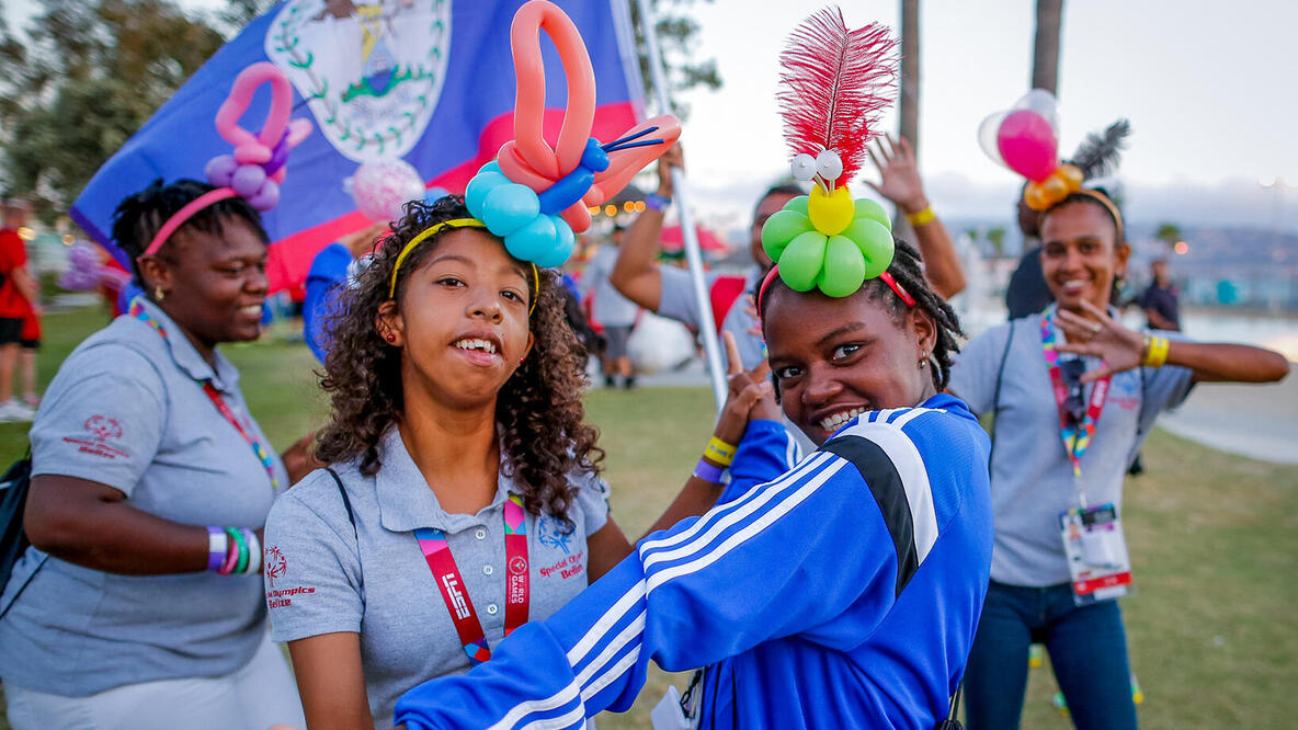 Vier Mädchen mit Ballons als Schmuck auf dem Kopf sind gut gelaunt und tanzen, im Hintergrund Fahnen der Special Olympics.