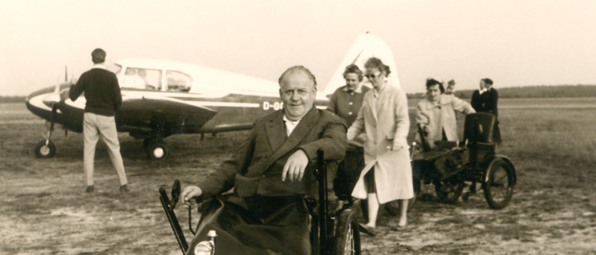 Bild eines Rollstuhlfahrers in den 60er Jahren - im Hintergrund ist ein kleines Flugzeug zu sehen.