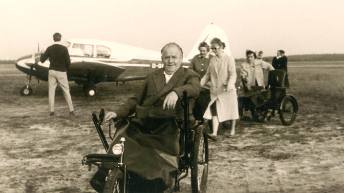 Bild eines Rollstuhlfahrers in den 60er Jahren - im Hintergrund ist ein kleines Flugzeug zu sehen.