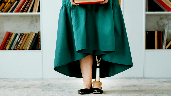 Foto: Die Beine einer Frau in smaragdgrünem Kleid vor einem weißen Bücherregal; das linke Bein der Frau ist eine Titanium-Prothese; die Frau hält mit durchgestreckten Armen einen Stapel Bücher vor der Hüfte