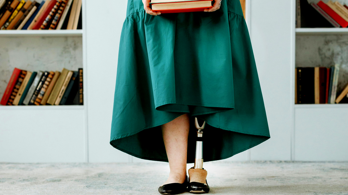 Foto: Die Beine einer Frau in smaragdgrünem Kleid vor einem weißen Bücherregal; das linke Bein der Frau ist eine Titanium-Prothese; die Frau hält mit durchgestreckten Armen einen Stapel Bücher vor der Hüfte