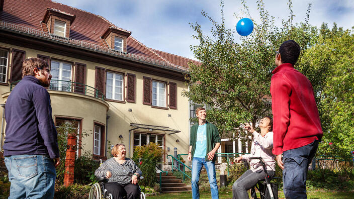 Fünf Personen, davon zwei im Rollstuhl, werfen sich vor der Villa Donnersmarck gegenseitig einen blauen Luftballon zu.