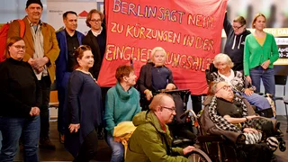 Eine Gruppe Menschen, darunter auch Rollstuhlfahrende, halten ein Banner mit der Aufschrift "Berlin sagt Nein zu Kprzungen in der Eingliederungshilfe" nach oben.
