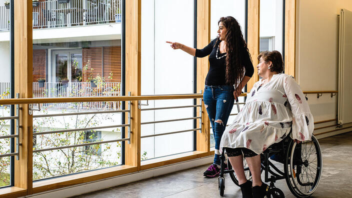 Zwei Frauen, eine davon im Rollstuhl, auf dem Flur des P.A.N. Zentrums. Die linke Frau zeigt mit dem Finger auf etwas draußen vor dem großen Panoramafenster.