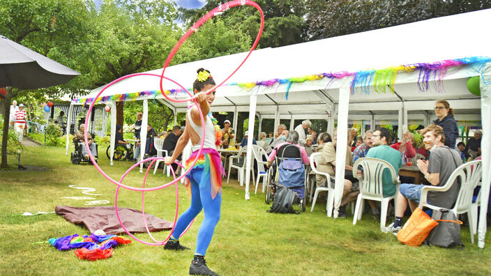 Foto: Sommerfest im Garten der Villa, viele Menschen sitzen unter weißen Gartenpavillons, im Vordergrund eine lachende Performance-Künstlerin mit Hula Hoop-Reifen