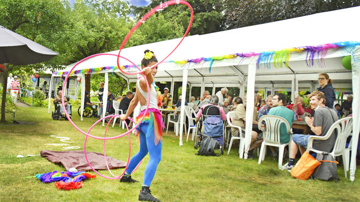 Foto: Sommerfest im Garten der Villa, viele Menschen sitzen unter weißen Gartenpavillons, im Vordergrund eine lachende Performance-Künstlerin mit Hula Hoop-Reifen