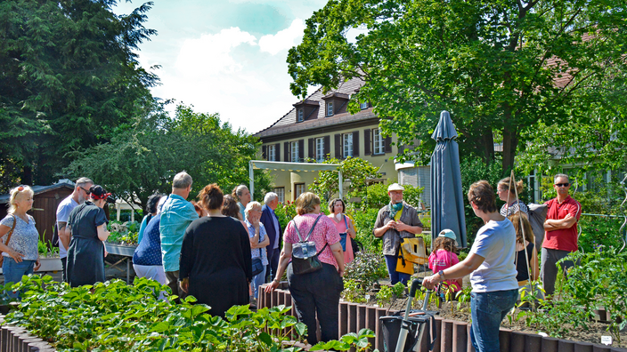 Foto: Eine bunt gemischte Menschengruppe im Hochbeetegarten der Villa Donnersmarck an einem Sonnentag; rechts im Vordergrund eine Dame am Rollator, im Hintergrund das Haus