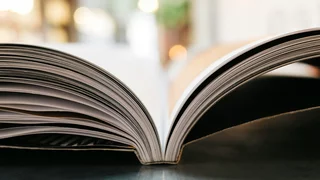 Ein aufgeschlagenes Buch, welches auf einem Tisch liegt wird von Sonnenlicht aus dem Hintergrund beleuchtet.