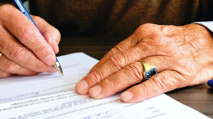 Die Hände einer älteren Person, die etwas auf einem Dokument unterschreibt.