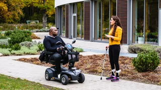 Person im Elektromobil unterhält sich auf einem Weg im Garten mit einer gehenden Person mit Gehstock vor dem P.A.N. Zentrum.