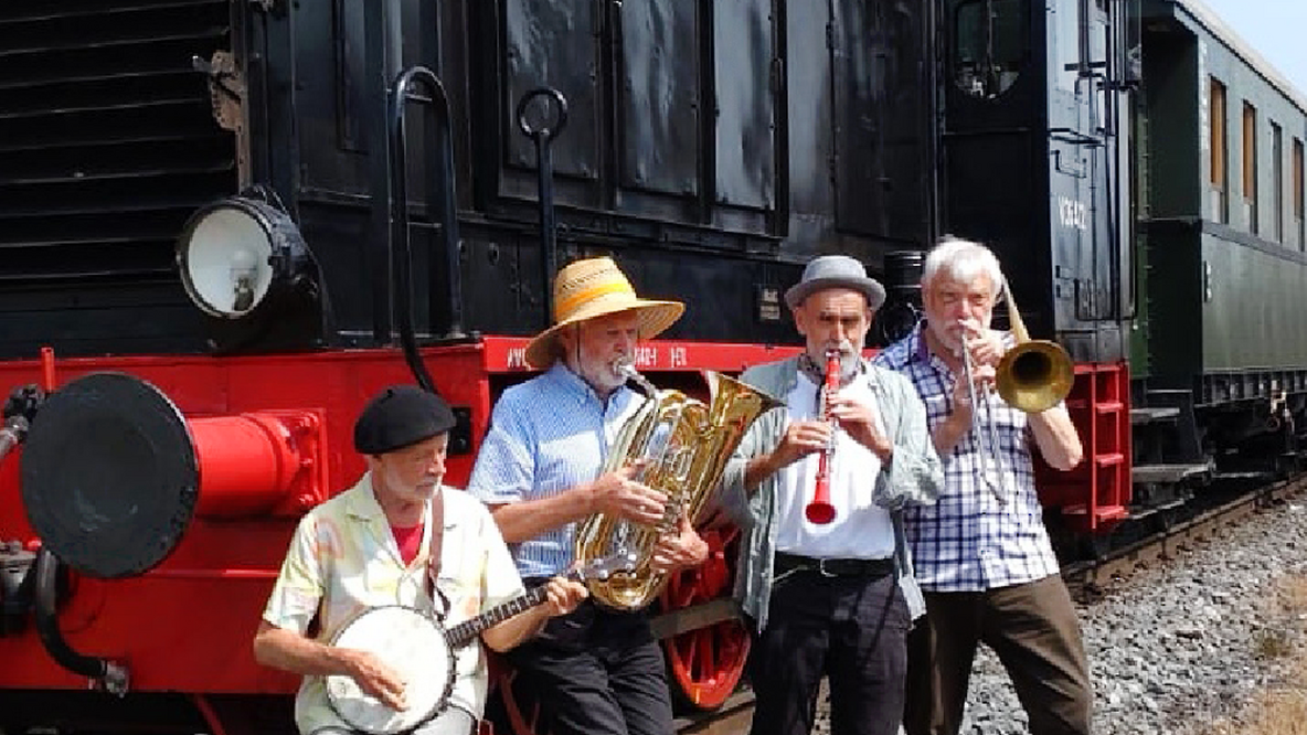 Foto: Die Band 5 o'clock International, vier weiße Männer reifen Alters mit ihren Instrumenten vor einer historischen Lokomotive