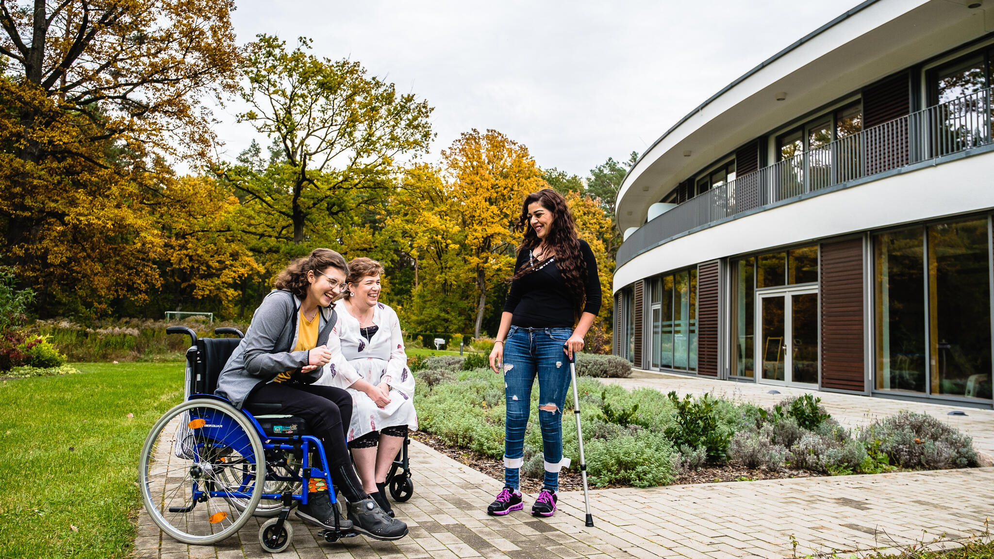 Drei Frauen, davon zwei im Rollstuhl und eine mit Gehhilfe, stehen gut gelaunt vor dem PAN Zentrum und unterhalten sich.