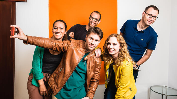 Zwei Mitarbeiterinnen und drei Mitarbeiter posieren lachend für ein Foto. Im Hintergrund ist eine orangene Fläche, der vorderste Mitarbeiter hält ein Smartphone zur Seite, um ein Selfie zu machen.