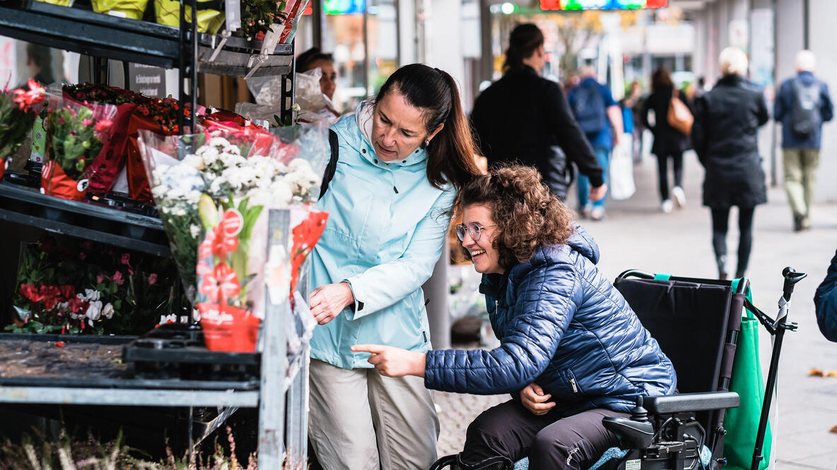 Zwei Frauen, davon eine im Rollstuhl, schauen sich die Angebote eines Blumenladens an.