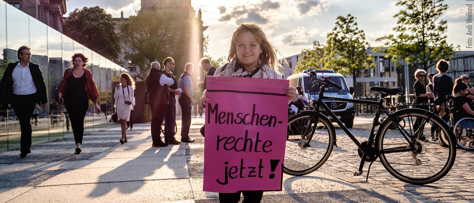 Eine kleinwüchsige Frau steht vor dem Reichstagsgebäude in Berlin und hält ein Plakat mit der Aufschrift "Menschen rechte jetzt!" hoch.