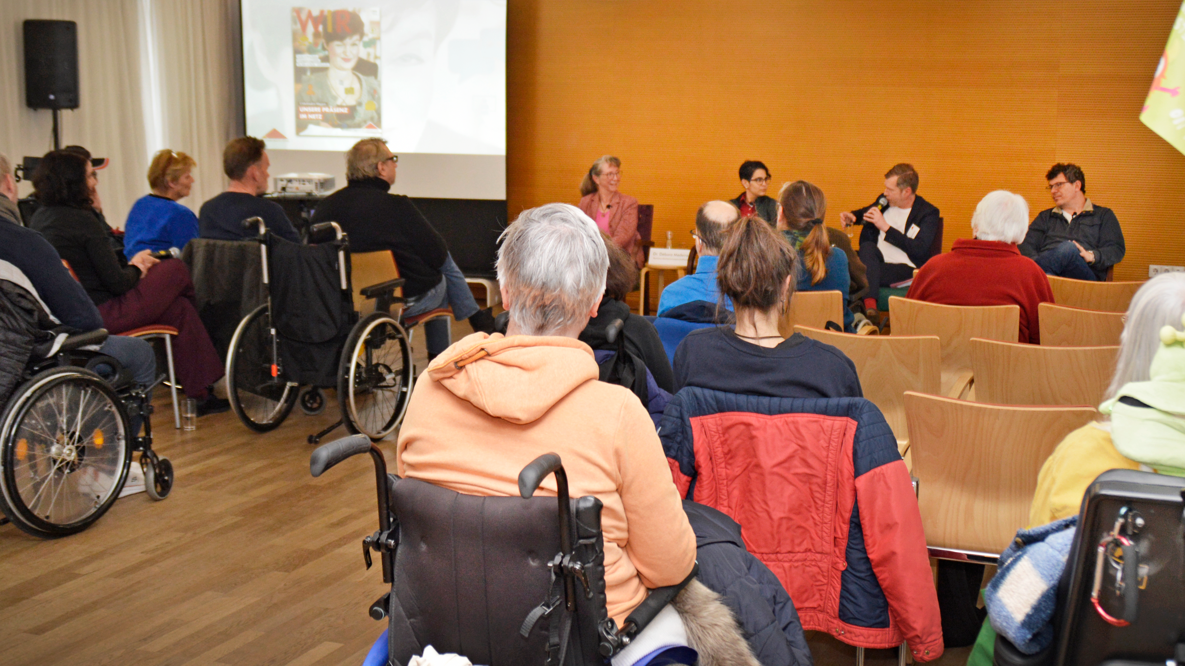 Foto: Blick aus dem Auditorium auf die Bühne bei der Dikussion in der Villa Donnersmarck; im Vordergund im Publikum zwei Damen im Rollstuhl