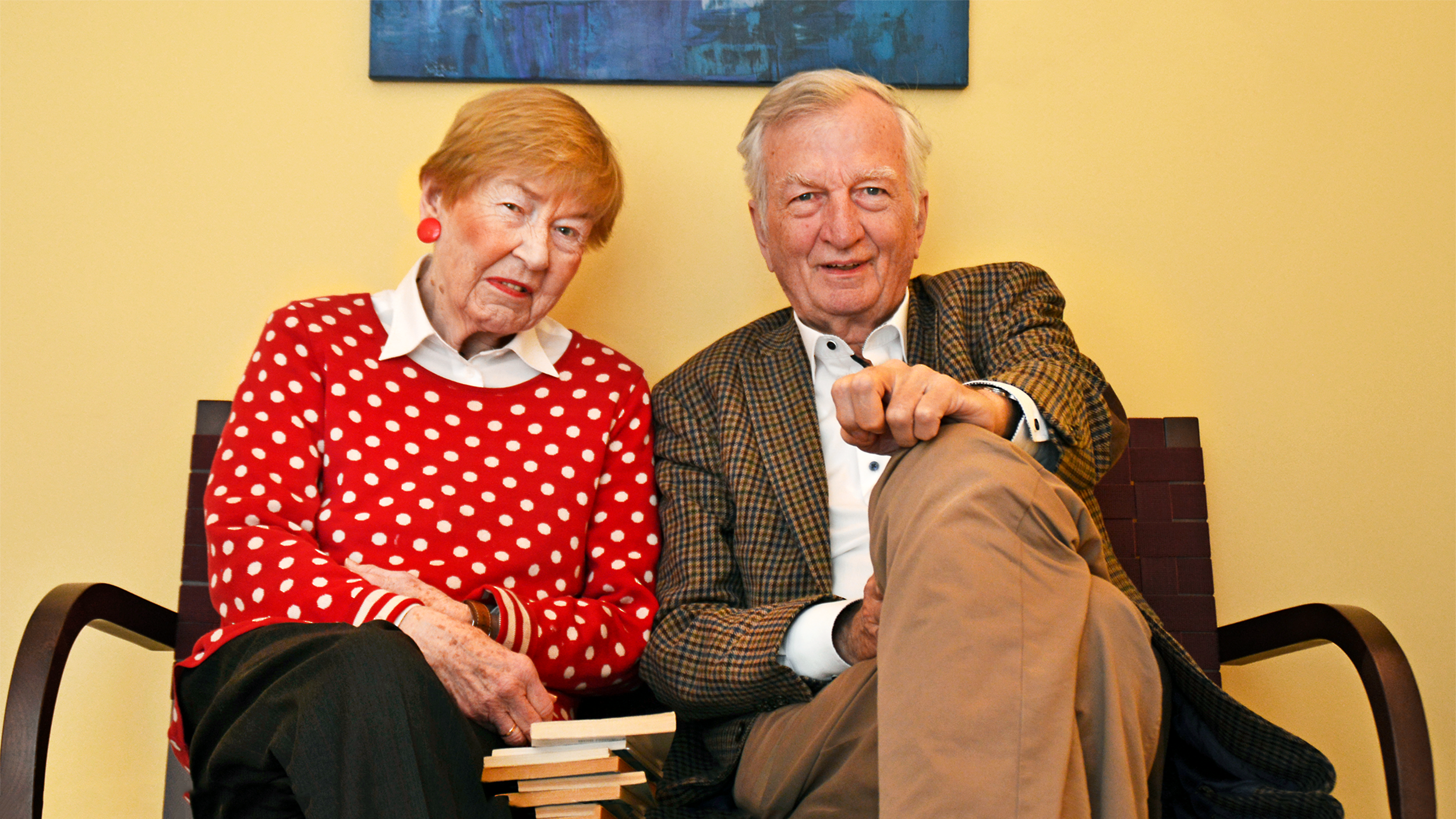 Foto: Hilke Dethlefs und Bernt Renzenbrink, ein Duo im fortgerückten Lebensalter, lächelnd auf einem Sofa sitzend vor einer gelben Wand